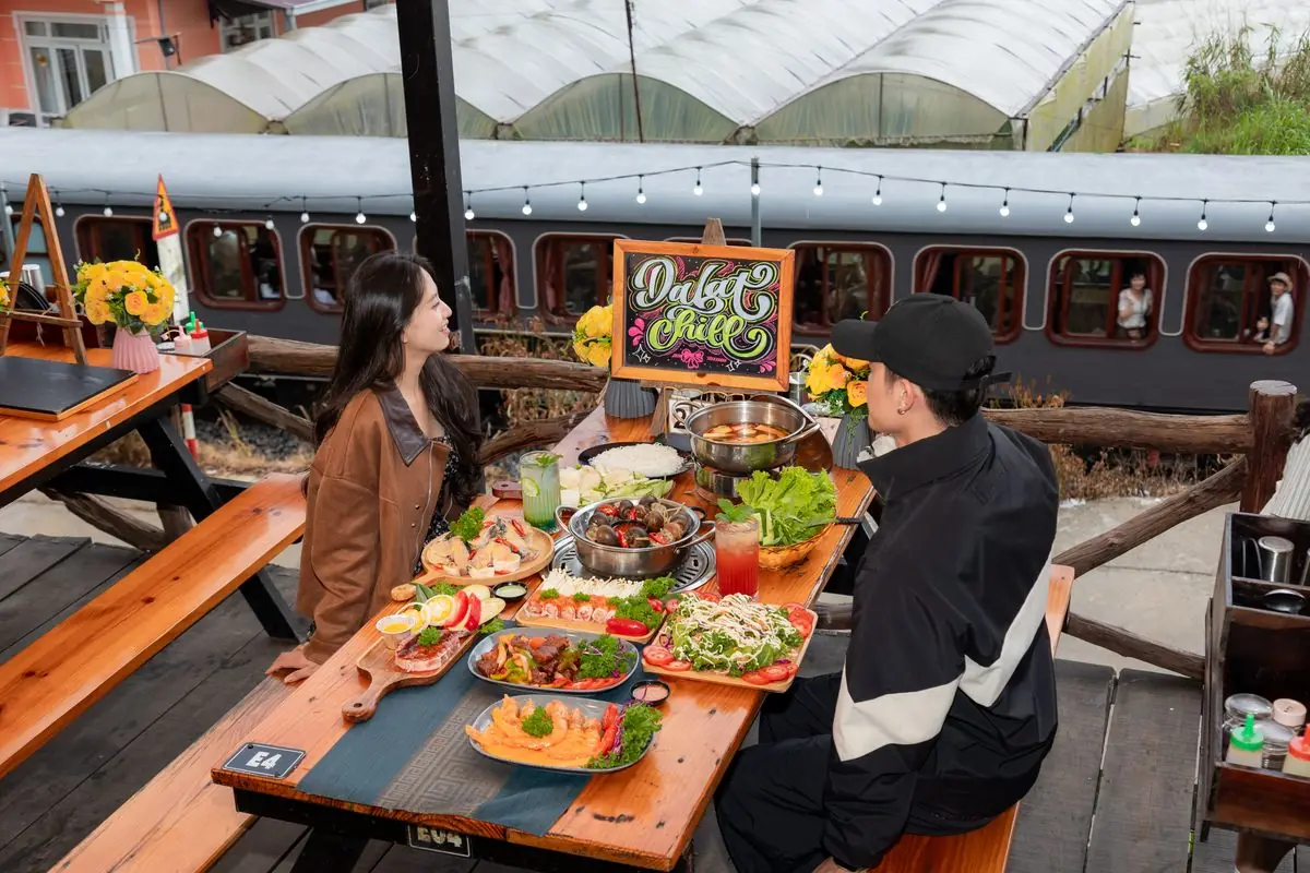 Cặp đôi ngồi bàn gỗ bày lẩu, khay thịt, hải sản và salad ở Trạm Dừng Chill, phía sau là toa tàu cửa sổ bo tròn của khu check-in trong quán