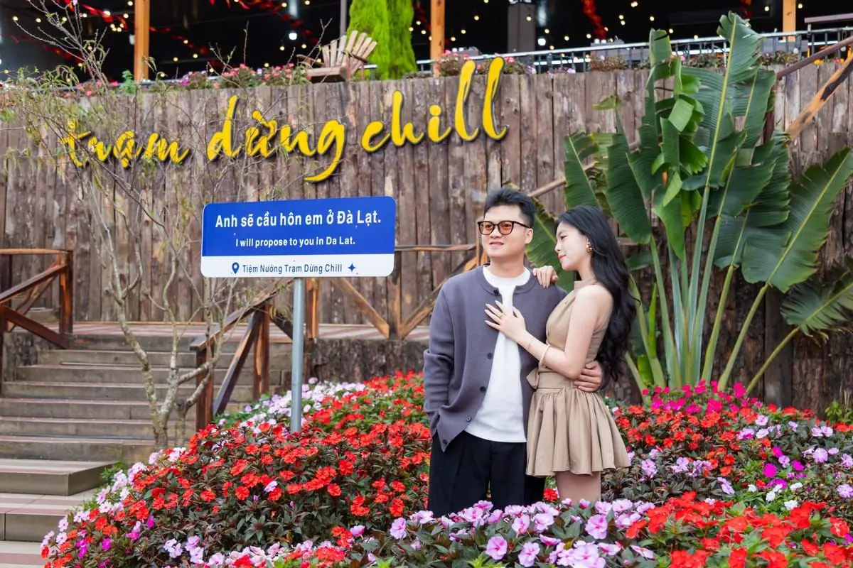 Cặp đôi đứng giữa vườn hoa cạnh biển “Anh sẽ cầu hôn em ở Đà Lạt”, phía sau là vách gỗ gắn chữ trạm dừng chill