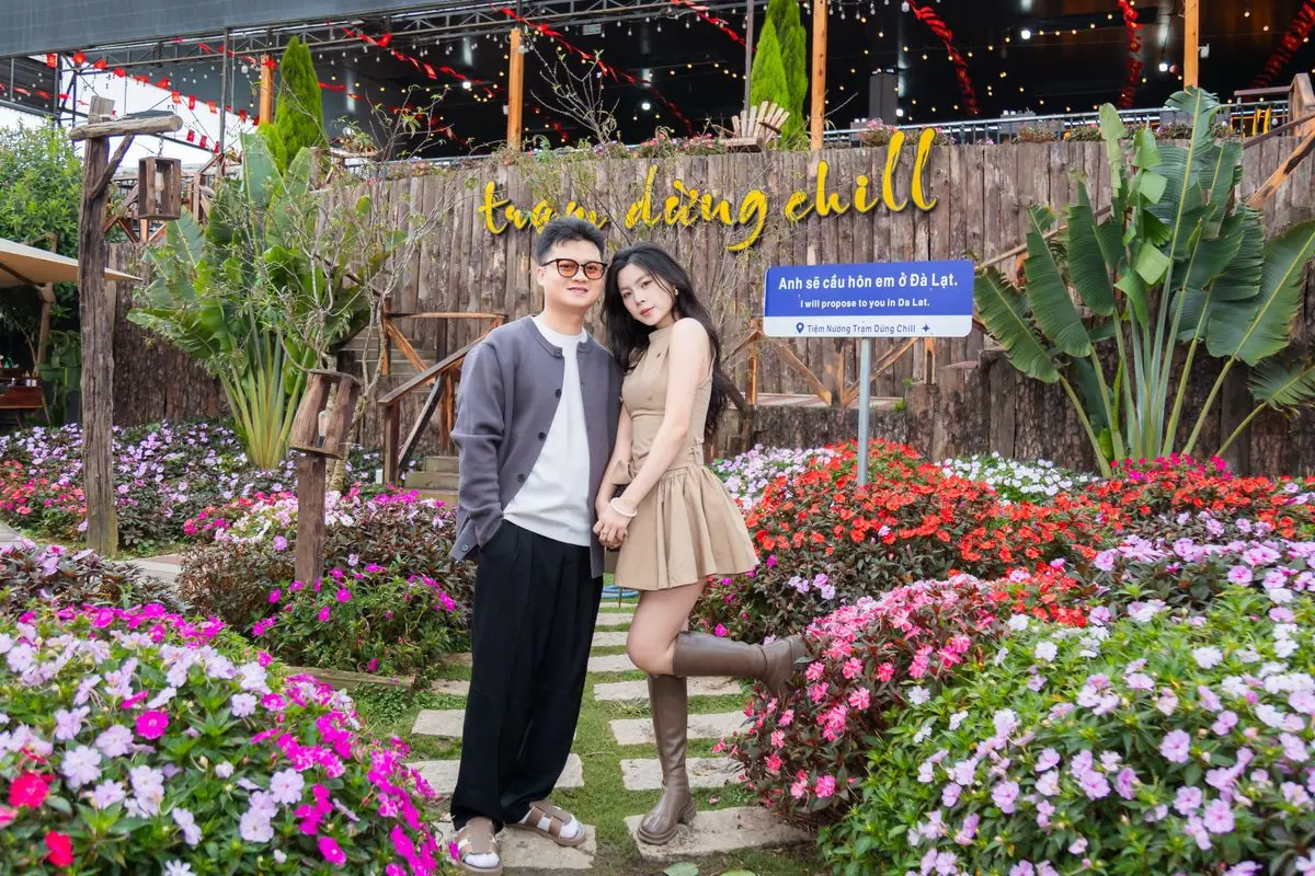 Cặp đôi đứng giữa vườn hoa trước vách gỗ gắn chữ trạm dừng chill, bên cạnh là biển chỉ đường “Anh sẽ cầu hôn em ở Đà Lạt”