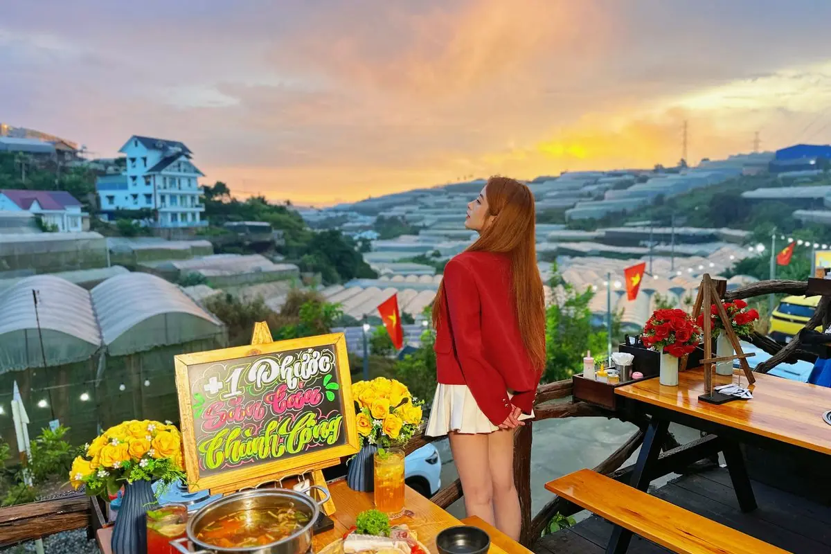 Cô gái áo đỏ đứng bên lan can ngắm hoàng hôn trên thung lũng nhà lồng, cạnh bàn ăn có bảng chữ viết tay và bình hoa vàng