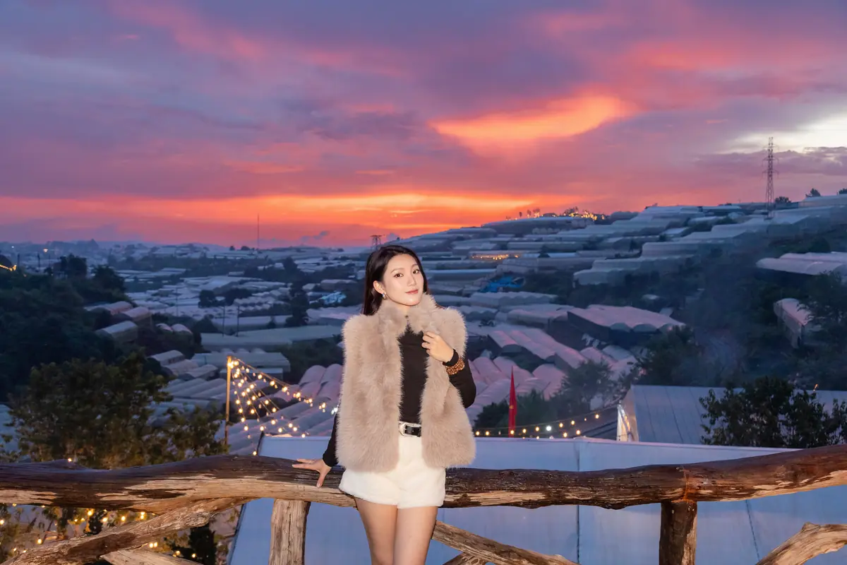 Cô gái mặc áo lông đứng tựa lan can gỗ ở Trạm Dừng Chill lúc hoàng hôn, dây đèn giăng phía sau, thung lũng nhà kính trải dài tới chân trời