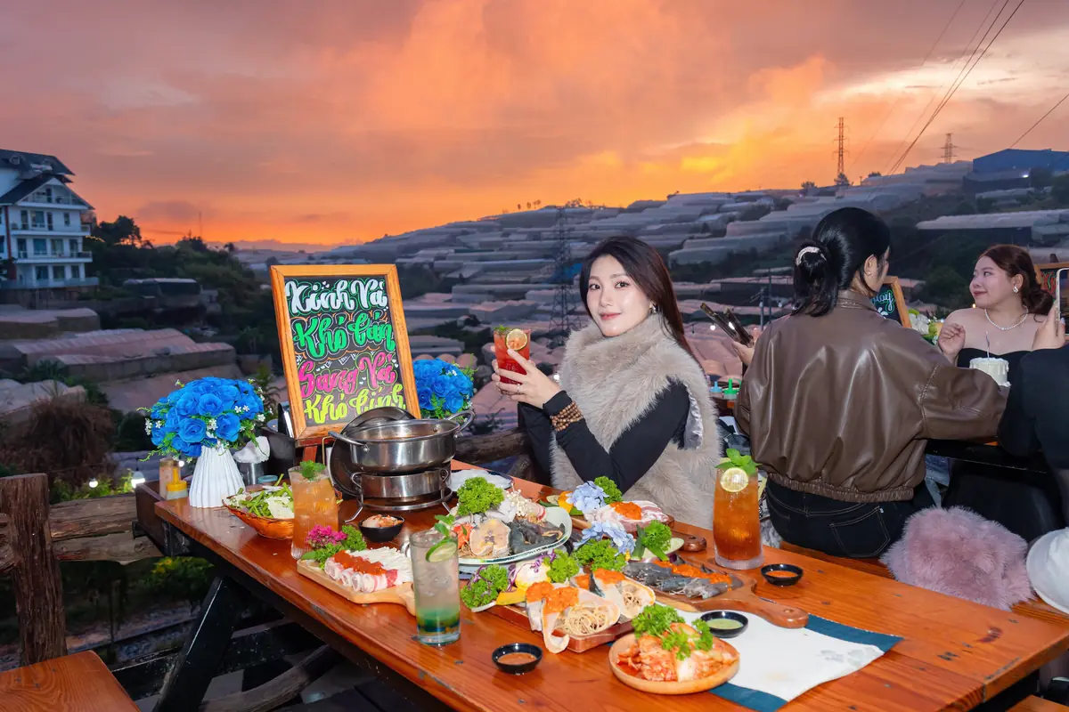 Cô gái mặc áo lông nâng ly bên nồi lẩu và khay hải sản ở Trạm Dừng Chill, trời hoàng hôn cam rực trên thung lũng nhà kính