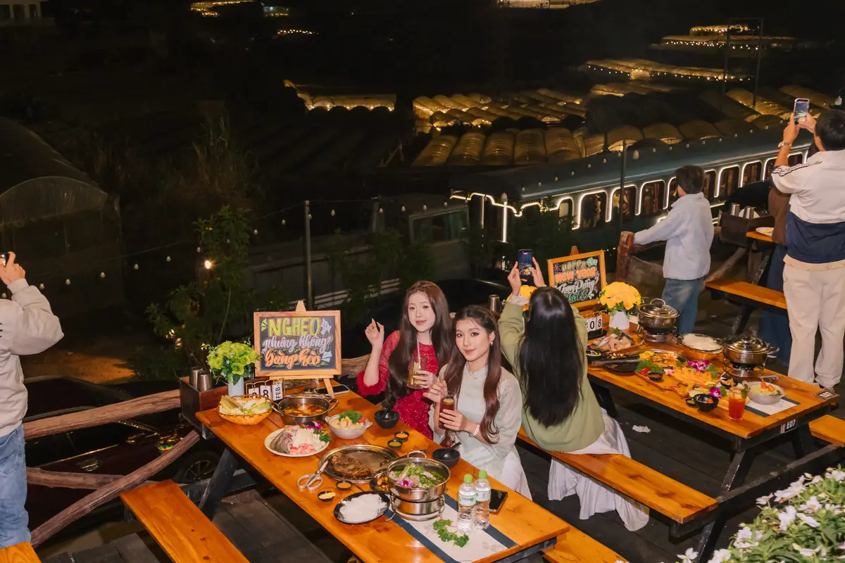 Nhóm bạn nâng ly bên bàn gỗ dài bày lẩu và đồ nướng ở Trạm Dừng Chill buổi tối, phía sau khách giơ điện thoại chụp toa tàu sáng đèn dưới lan can, xa hơn là nhà lồng lên đèn