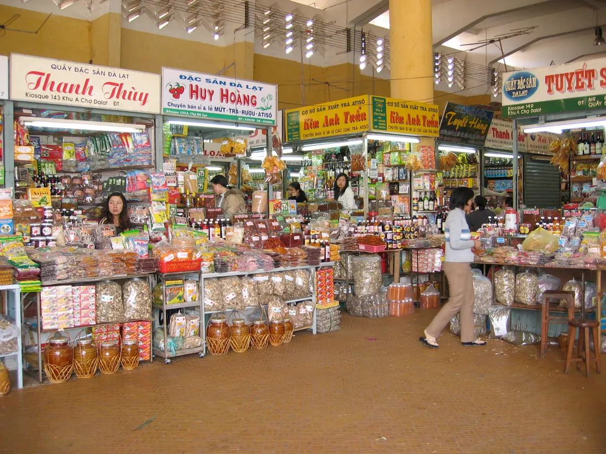Dãy quầy đặc sản trong chợ Đà Lạt bày kín mứt, trà, rượu và hũ ngâm, biển hiệu ghi mứt, trà, rượu, atiso