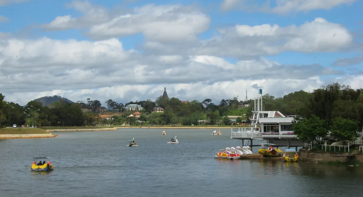 Pedal boats on Xuan Huong Lake in central Da Lat, with pine-covered hills under a cloudy blue sky