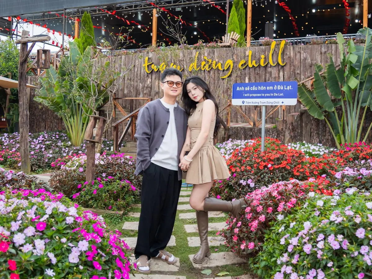 Cặp đôi đứng giữa vườn hoa trước vách gỗ gắn chữ trạm dừng chill, bên cạnh là biển chỉ đường “Anh sẽ cầu hôn em ở Đà Lạt”