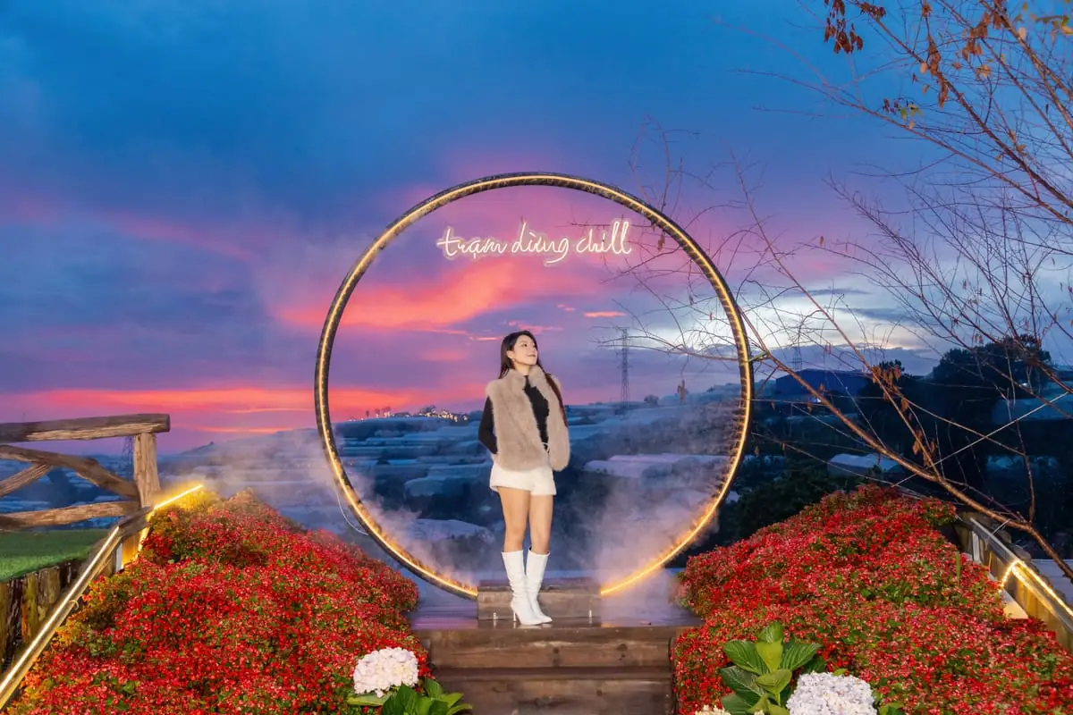 Cô gái đứng giữa cổng tròn gắn chữ đèn trạm dừng chill, hai bên là luống hoa đỏ và cẩm tú cầu, trời hoàng hôn tím hồng phía sau