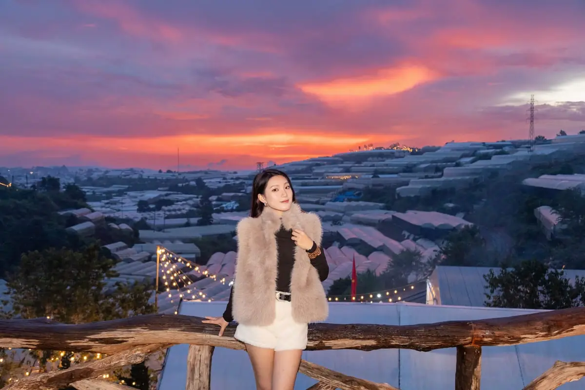 Cô gái mặc áo lông đứng tựa lan can gỗ ở Trạm Dừng Chill lúc hoàng hôn, dây đèn giăng phía sau, thung lũng nhà kính trải dài tới chân trời
