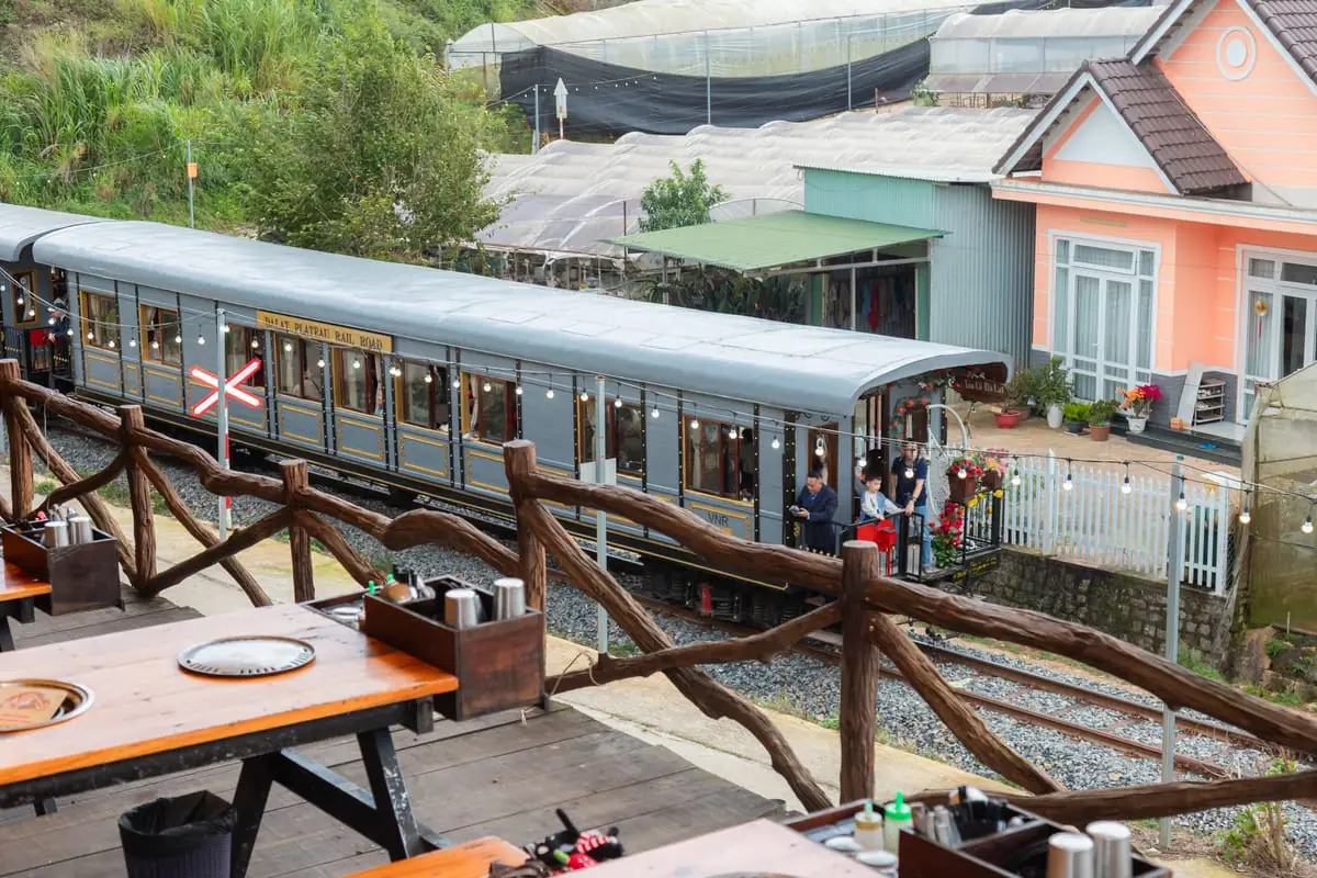Toa tàu xám Dalat Plateau Rail Road trên đường ray ngay dưới lan can gỗ của quán, có biển báo giao cắt đường sắt, phía sau là nhà kính và căn nhà hồng