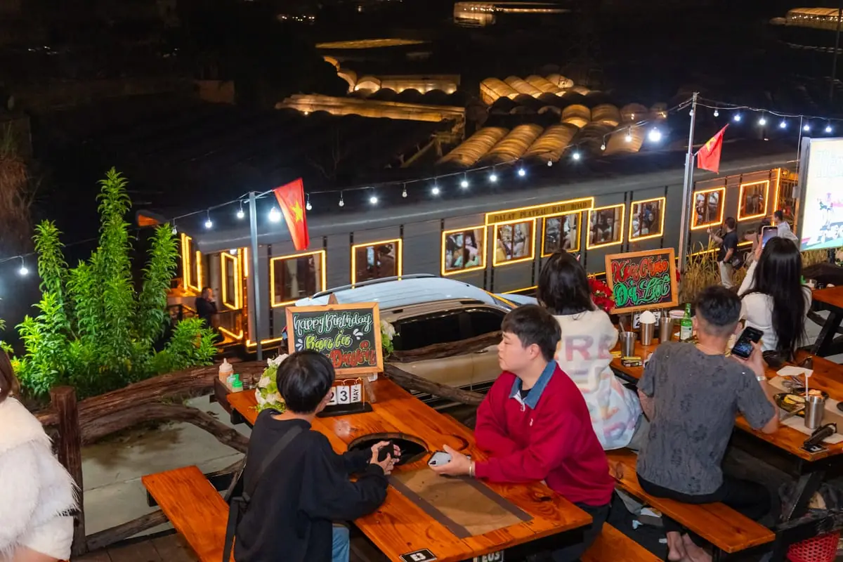 Khách ngồi bàn gỗ bên lan can buổi tối, ngay dưới là toa tàu Dalat Plateau Rail Road viền đèn vàng, xa hơn là mái nhà lồng sáng đèn trong thung lũng